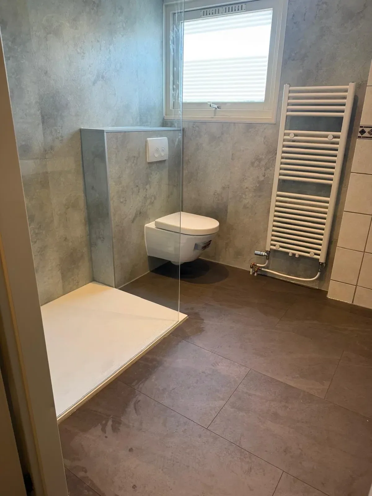 Badkamer met hangtoilet, grijze wandpanelen, douchebak en witte handdoekradiator onder het raam