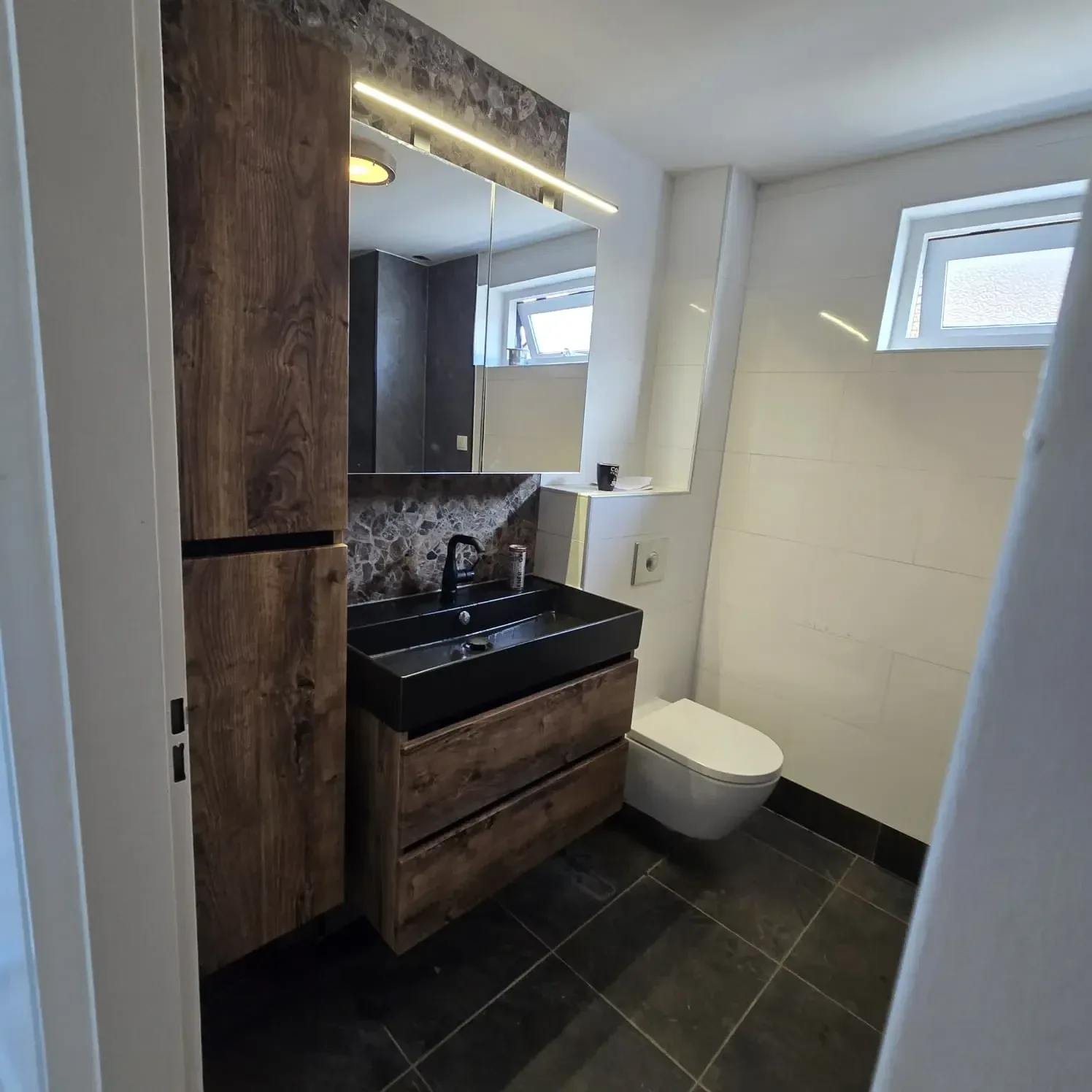 Badkamer met houtlook hoge kast, zwarte wastafel op houten meubel, spiegelkast met LED en hangtoilet