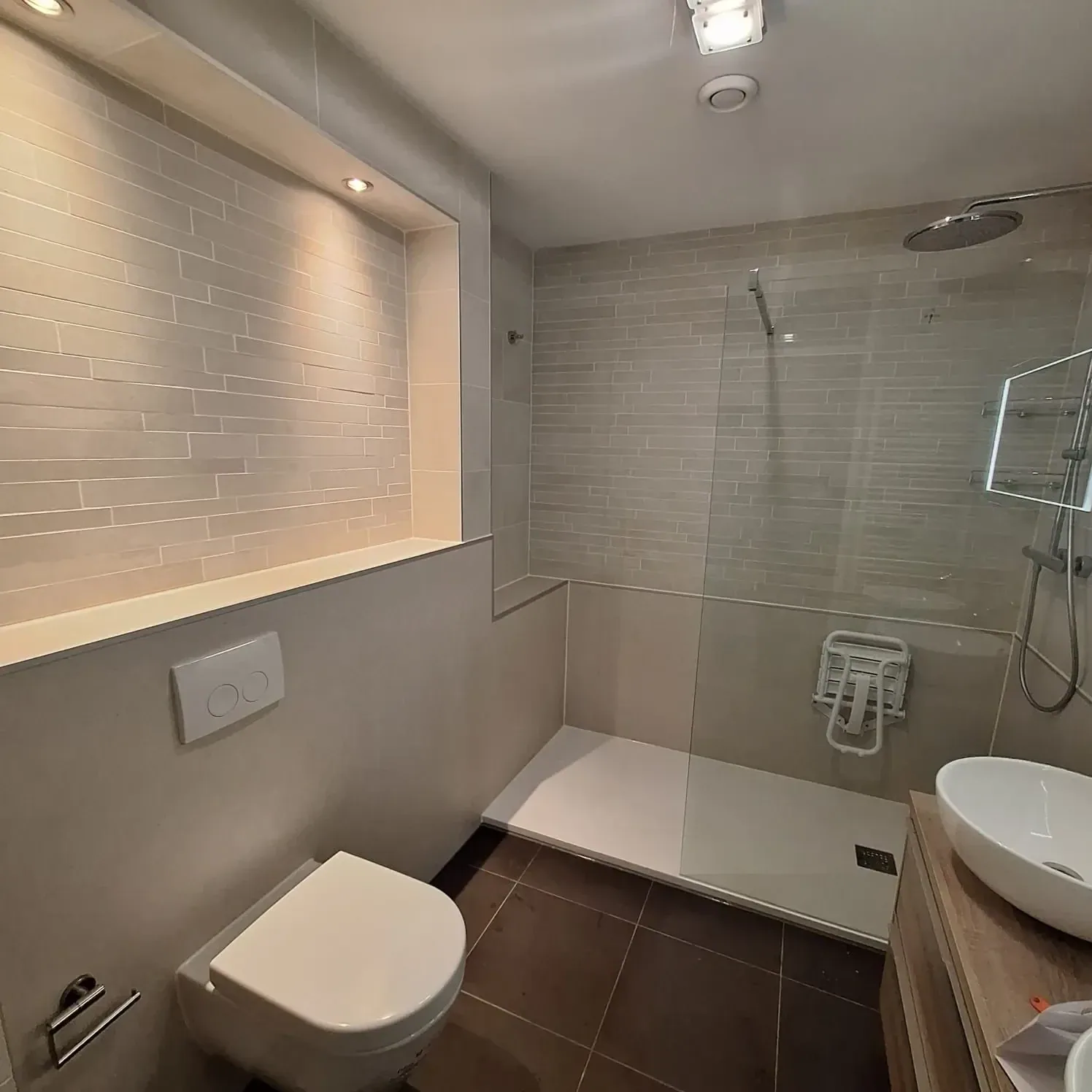 Badkamer met inloopdouche met douchezitje, hangtoilet, nis met verlichting en waskom op houten meubel