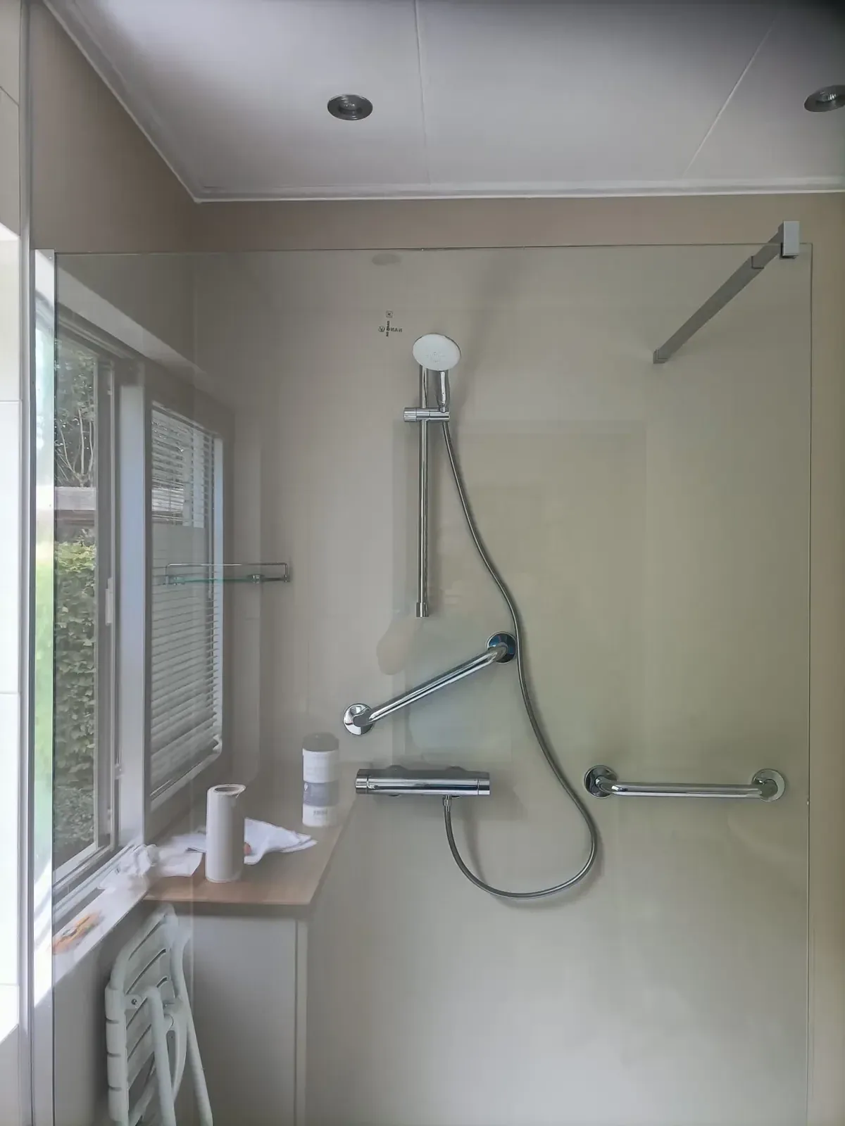 Douche bij raam met glazen wand, handdouche op glijstang, twee steunbeugels en opklapbaar zitje