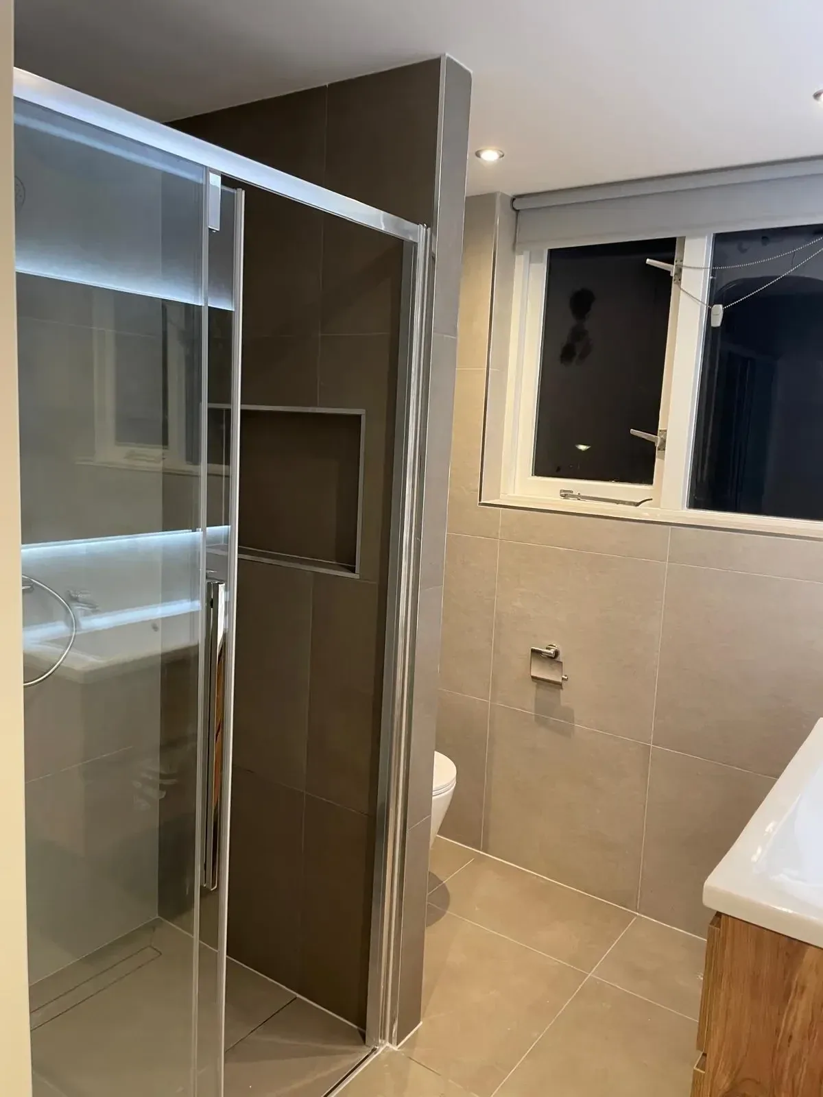 Douche met glazen wand en verlichte nis, beige tegels, raam en houten wastafelmeubel