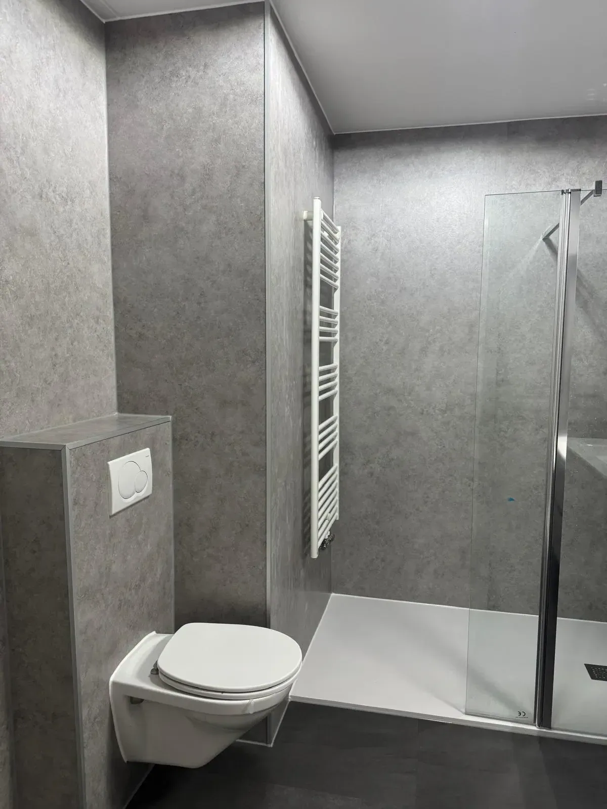 Badkamer met grijze betonlook wandpanelen, hangtoilet op voorzetwand en inloopdouche met glazen wand