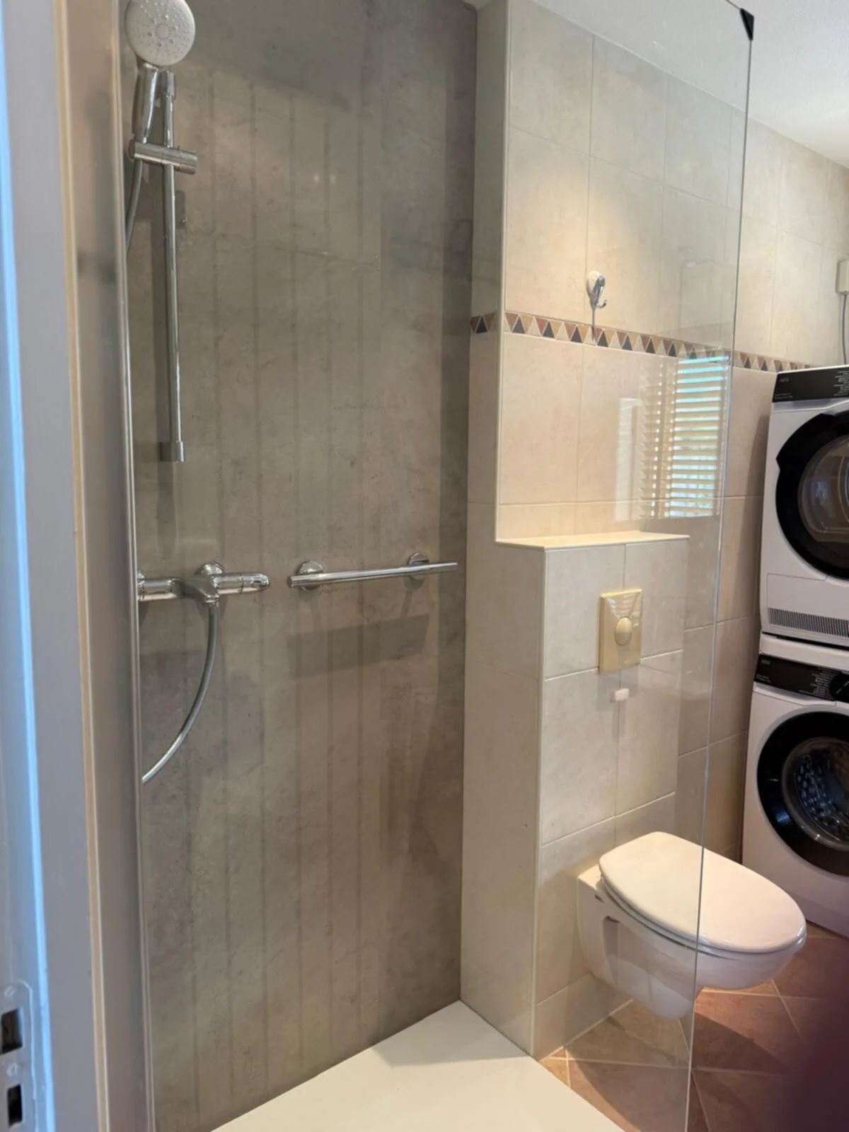 Inloopdouche met grijze betonlook wandpanelen, glazen wand en beugel, ernaast hangtoilet en wasmachine