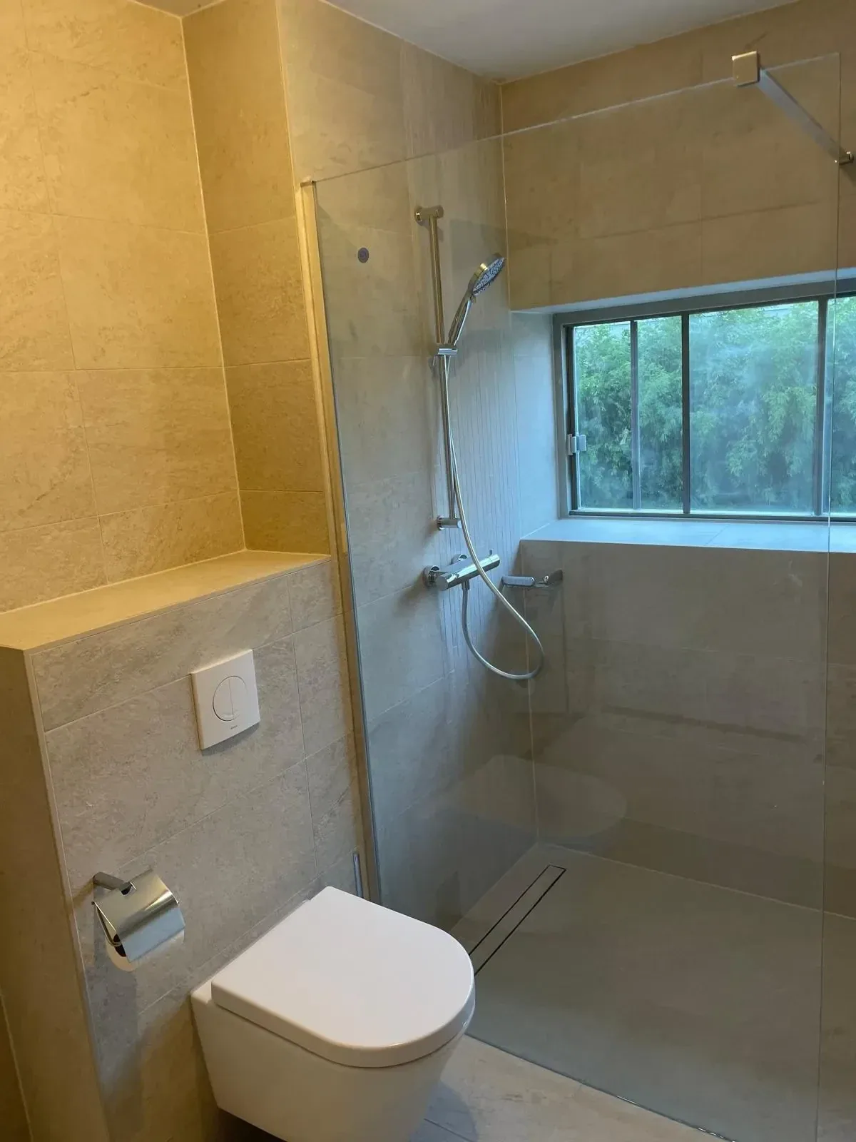 Inloopdouche met glazen wand en douchegoot onder raam, ernaast hangtoilet tegen beige betegelde voorzetwand