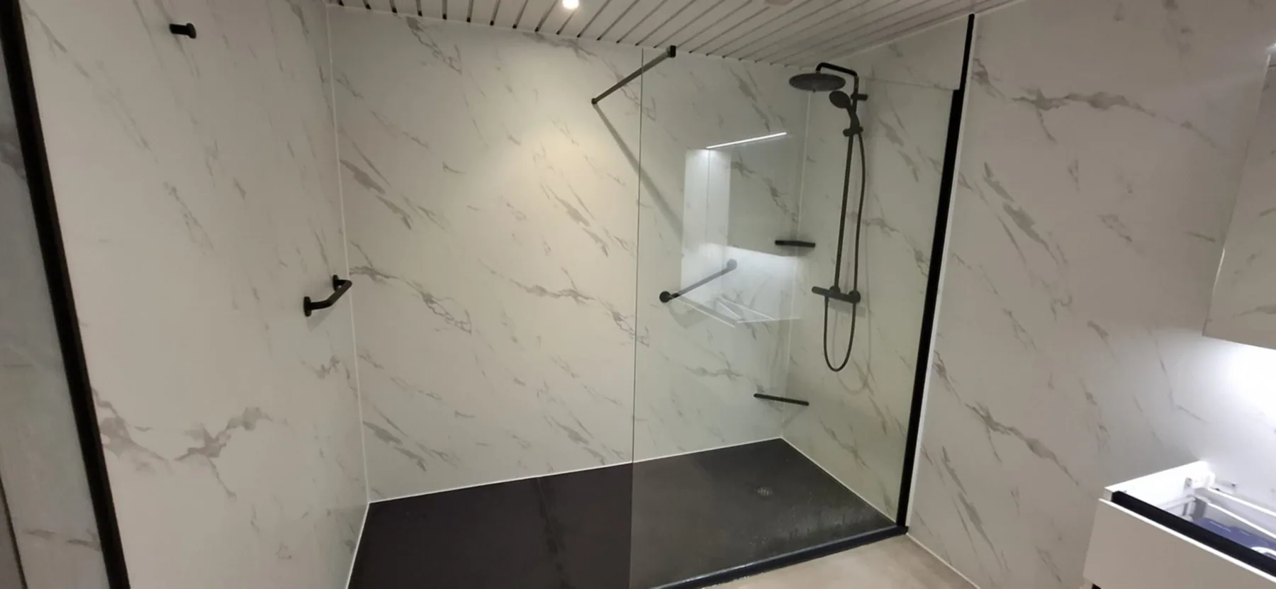 Ruime inloopdouche met witte marmerlook wandpanelen, zwarte regendouche, nis met verlichting en steunbeugels