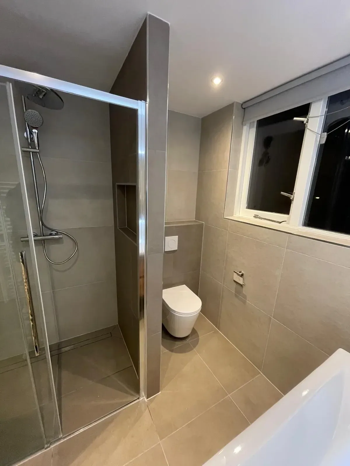 Inloopdouche met glazen wand en nis, ernaast hangtoilet en ligbad in badkamer met grote beige tegels