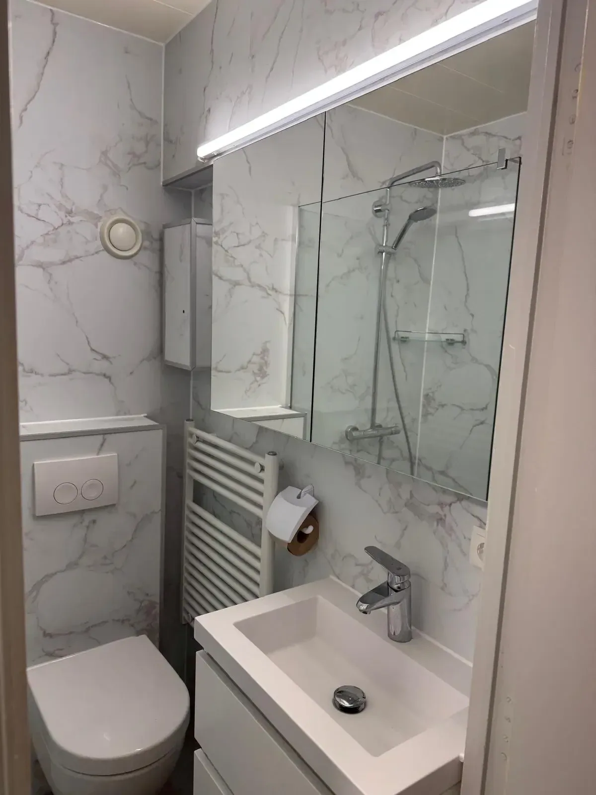 Kleine badkamer met witte marmerlook panelen, hangtoilet, wastafel, radiator en douche met glazen wand
