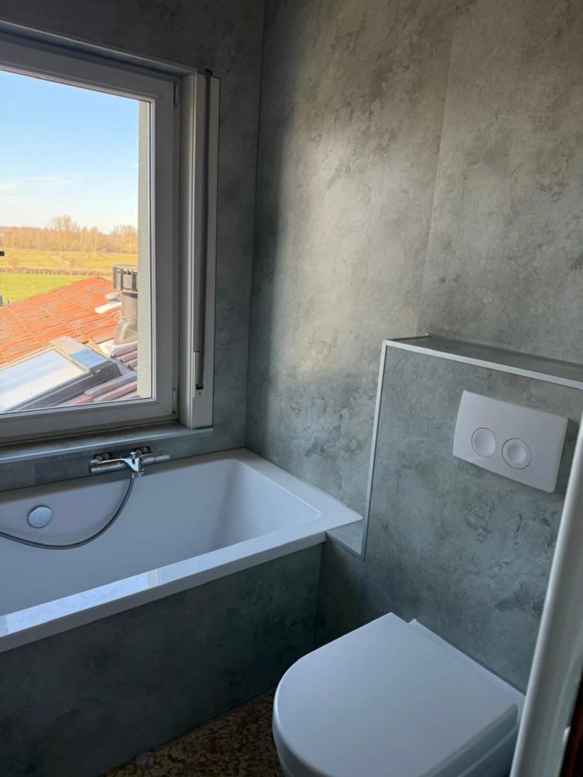 Ligbad onder het raam met uitzicht, wanden in grijze betonlook panelen en hangtoilet met bedieningsplaat