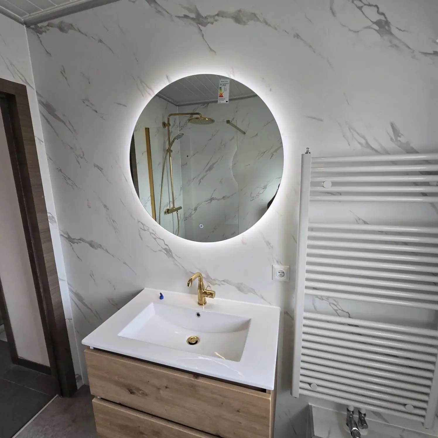 Ronde ledspiegel boven eiken wastafelmeubel met gouden kraan, marmerlook wandpanelen
