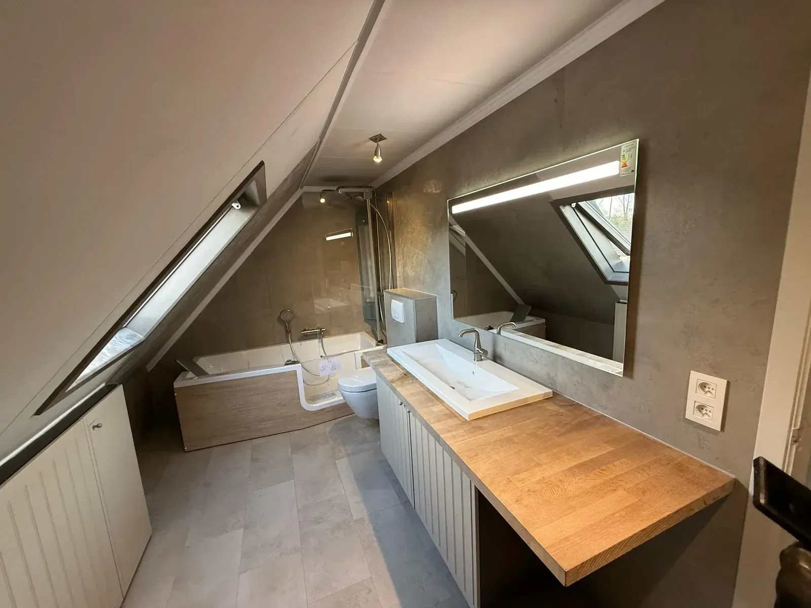 Zolderbadkamer met grijze betonlook wandpanelen, ligbad met instapdeur, hangtoilet, wastafel op houten blad en LED-spiegel